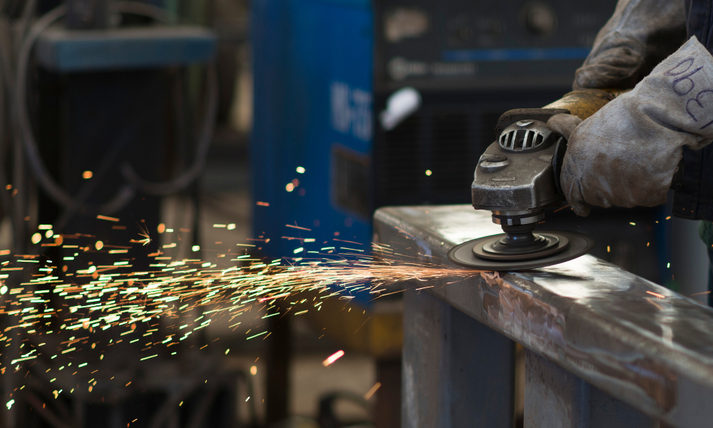 A close-up action shot of a person wearing protective gloves and a face shield using an angle grinder to cut a metal beam, with bright sparks flying in the foreground.